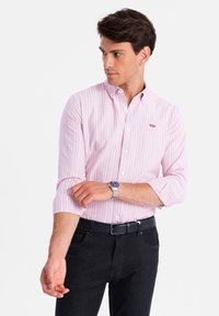 Wit en roze gestreept overhemd met kraag en opgerolde mouwen. Zwarte riem en donkere spijkerbroek zichtbaar. Zilveren horloge om de pols.
