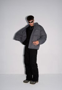 Szara, ocieplana kurtka z kapturem, zapinana na zamek, z wewnętrznym wzorem, noszona na czarnej bluzce i czarnych spodniach cargo. Szare obuwie i okulary przeciwsłoneczne.