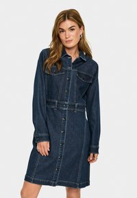Frau mit langen Haaren, die ein dunkelblaues, knopfbares Jeanskleid mit langen Ärmeln und Brusttaschen trägt, steht vor einem schlichten Hintergrund.