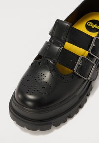 Czarne skórzane buty z podwójnymi klamrami, perforowanymi detalami, masywną podeszwą i jaskrawym żółtym wnętrzem. Okrągły nos i teksturowana powierzchnia.