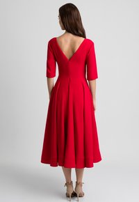 Kobieta z długimi brązowymi włosami, ubrana w czerwoną sukienkę midi z głębokim dekoltem na plecach oraz rękawami do łokci, do której dobrała beżowe sandały na wysokim obcasie.