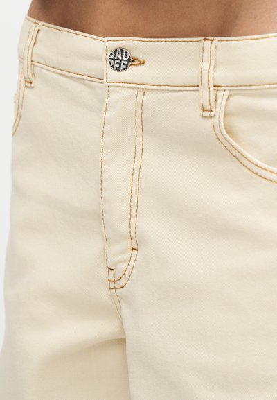 Εικόνα κοντινού πλάνου της περιοχής της μέσης με μπεζ παντελόνι από τζιν ύφασμα, με εμφανή ραφή, κουμπί και μπροστινές τσέπες.