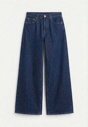 Tummansiniset leveälahkeiset denimfarkut, joissa etuosassa nappi ja vetoketju, vyölenkit ja viiden taskun malli yksivärisellä vaalealla taustalla.