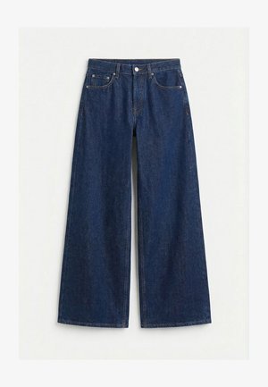 Tummansiniset leveälahkeiset denimfarkut, joissa etuosassa nappi ja vetoketju, vyölenkit ja viiden taskun malli yksivärisellä vaalealla taustalla.