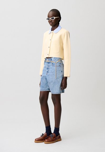 Giacca gialla a taglio corto con due tasche e trama; camicia blu con colletto sotto. Pantaloncini di denim azzurro con dettaglio a catena. Scarpe marroni con lacci.
