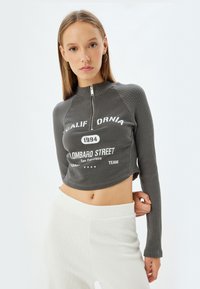 Gray cropped sweatshirt with ribbed texture, high neckline, and front zipper. Features white printed text and a relaxed fit.