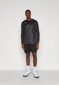 Schwarzes langärmeliges Sportshirt mit einem geometrischen Muster, kombiniert mit schwarzen Shorts und weißen Socken. Das Outfit ist mit Puma-Branding versehen.