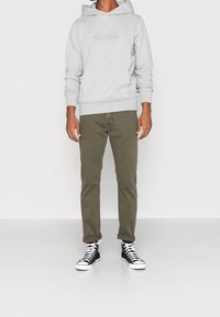Felpa grigia con tasca frontale, caratterizzata da un piccolo logo. Pantaloni slim-fit verde oliva e sneakers alte nere con suola bianca.