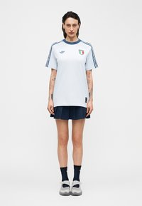 Femme debout, face à l'avant, portant un maillot de football italien blanc et bleu, une jupe bleu marine, des chaussettes noires et des baskets blanches.