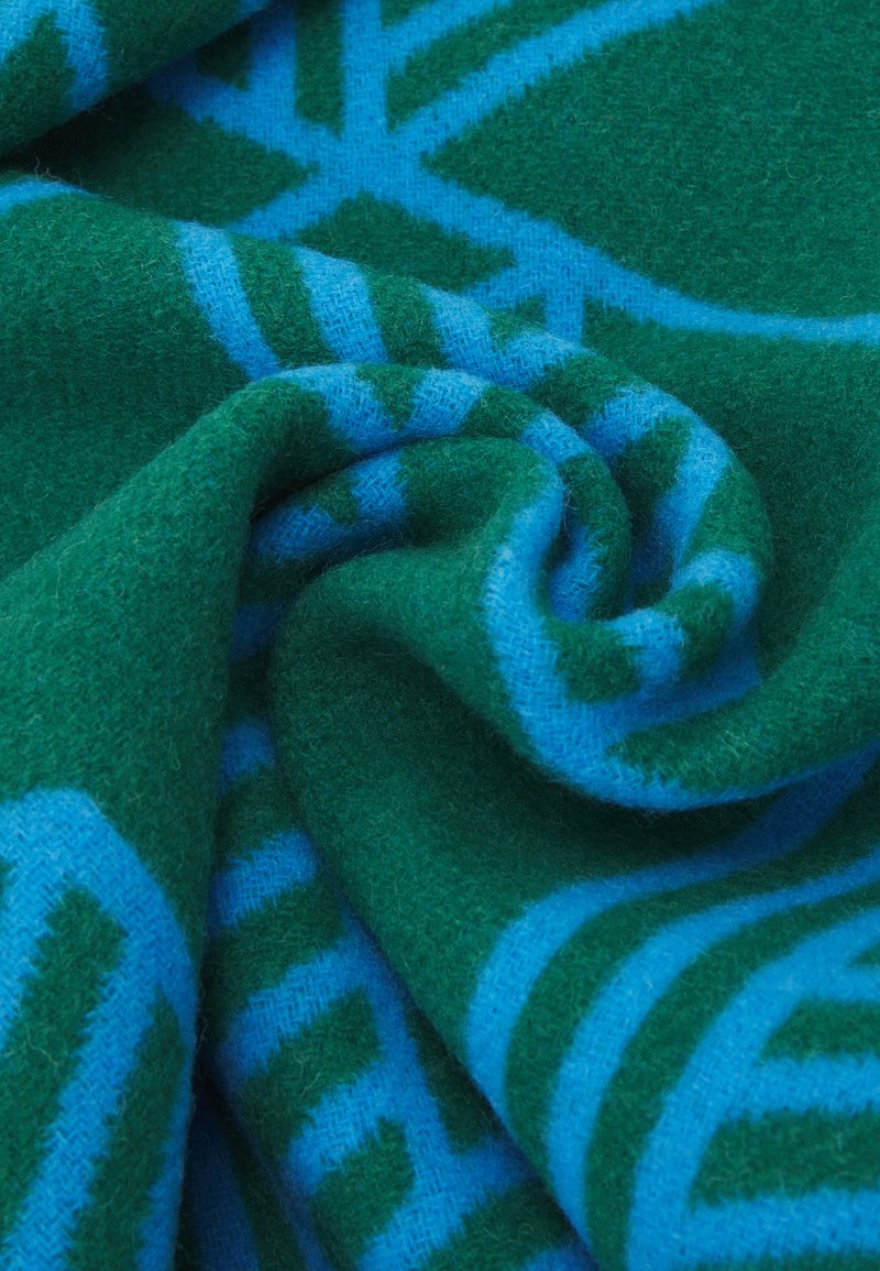 Manta de lana con una textura suave, predominantemente verde con patrones y rayas geométricas azules, mostrando un borde rizado.