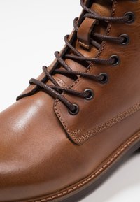 Hnědé kožené šněrovací boty s hladkým povrchem, kovovými očky a vyztuženým šitím podél podešve pro odolnost.
