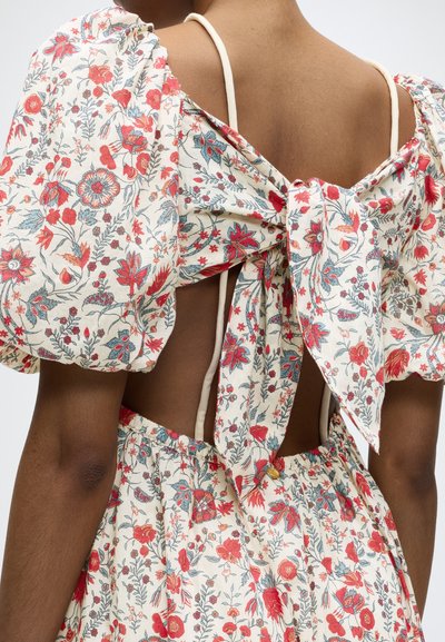 Vue de dos d'une personne portant une robe crème avec un motif floral rouge et bleu, manches bouffantes, dos ouvert, nœud noué et fines bretelles blanches.