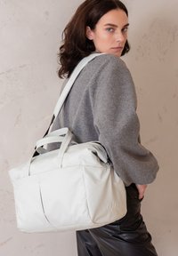 Borsa multifunzionale grigia chiara con doppi manici e tracolla regolabile, caratterizzata da una superficie liscia e raffinata e un branding sottile.