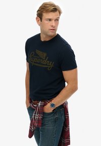 Camiseta de algodón azul marino con un logo dorado de "Superdry", combinada con jeans azules y una camisa de franela a cuadros rojos atada a la cintura.