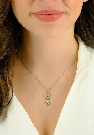 Collana d'argento con ciondolo a forma di fiocco di neve, design intricato e catena sottile. Indossata sopra una camicetta bianca con scollo a V.