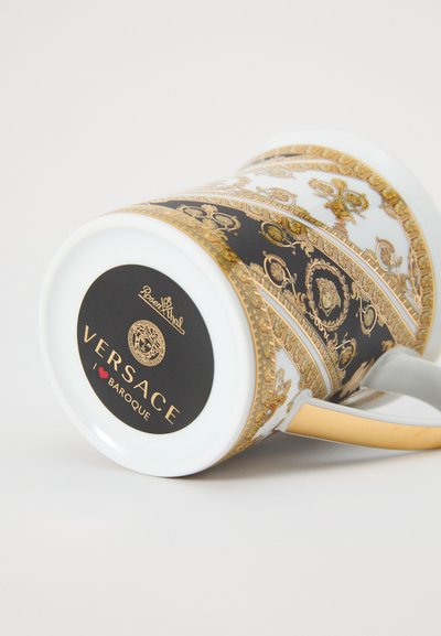 Κούπα Versace με λευκή βάση, μαύρα και χρυσά μπαρόκ σχέδια, χρυσή λαβή και στρογγυλό λογότυπο στο κάτω μέρος.