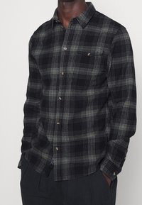 Black and green checkered flannel shirt with a button-down collar, front pocket, and brown buttons. Soft texture and long sleeves.