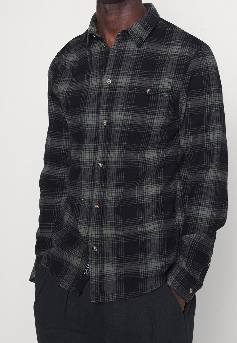 Black and green checkered flannel shirt with a button-down collar, front pocket, and brown buttons. Soft texture and long sleeves.