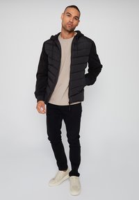 Sort pufferjakke med en tekstureret front og glatte ærmer, parret med en lys beige skjorte, sorte tætsiddende jeans og cremede sneakers.