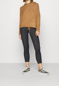 Felpa con cappuccio a maniche lunghe di colore marrone chiaro e texture, con coulisse, abbinata a leggings in denim scuro e scarpe verdi con dettagli bianchi.