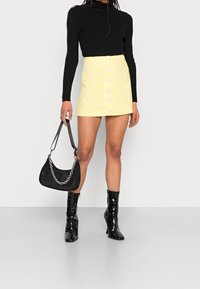 Mini jupe jaune avec boutons sur le devant, haut noir à manches longues côtelé, bottes noires vernies jusqu'au genou et sac à épaule noir avec une bandoulière en chaîne.