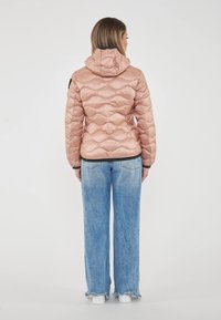 Donna in piedi con le spalle rivolte, indossa una giacca trapuntata rosa con cappuccio, jeans a gamba larga azzurri con orli sfilacciati e sneakers bianche.