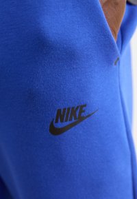 Nike apģērbs košā zilā audumā, ar gludu tekstūru un melnu logotipa akcentu, izcili parādīts sānos.