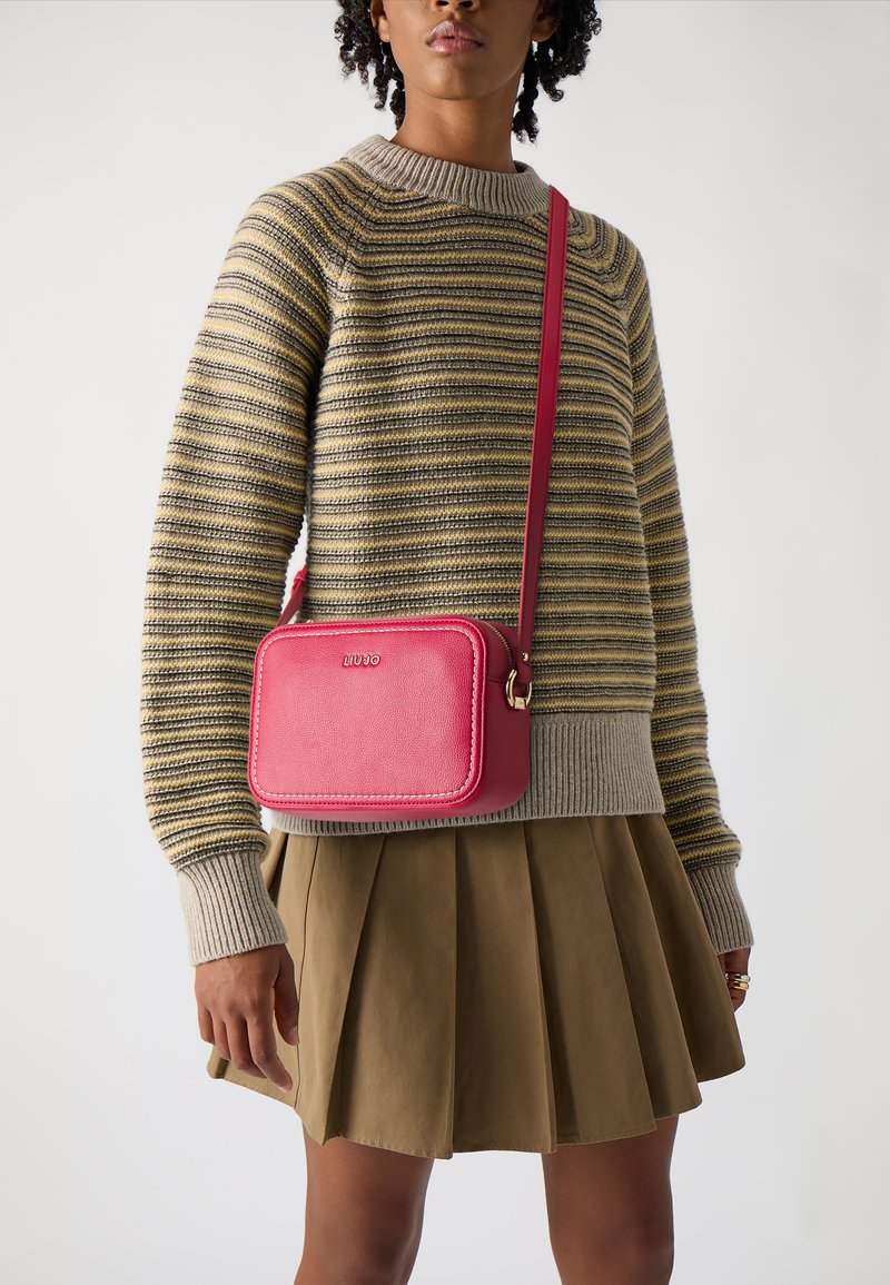 Borsa a tracolla in pelle rosa con forma rettangolare, dettagli in oro e logo. Indossata con un maglione a righe e una gonna plissettata.