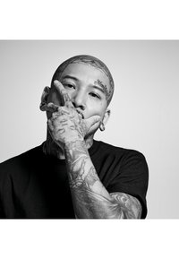 Porträtt av en person med rakad skalle och omfattande tatueringar, iförd en svart t-shirt. Kännetecken inkluderar ansiktspiercingar och synliga tatueringar.