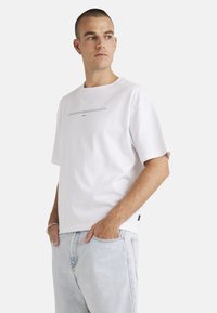 White cotton t-shirt with a crew neck and short sleeves. Features a black printed text design across the chest. Paired with light blue jeans.