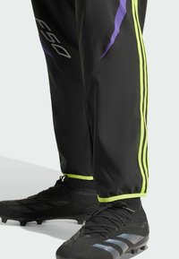 Czarne spodnie sportowe z purpurowymi i neonowozielonymi akcentami, z trzema pionowymi paskami po bokach. W połączeniu z czarnymi korkami.