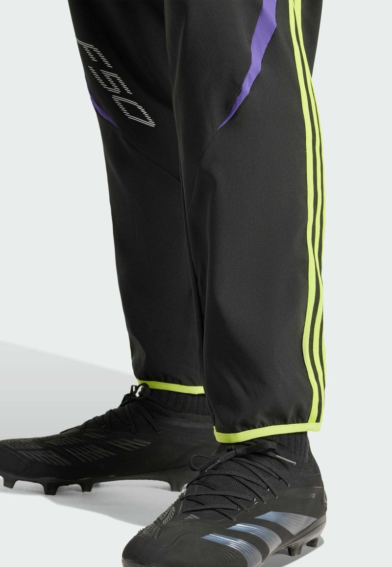 Pantalones deportivos negros con acentos en púrpura y verde neón, presentando tres rayas verticales a lo largo de los lados. Combinados con zapatillas negras.