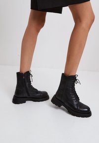 Sorte snørebånd ankelstøvler lavet af tekstureret læder med en chunky sål og lynlåsdetalje på siden; har rund tå og syede detaljer.