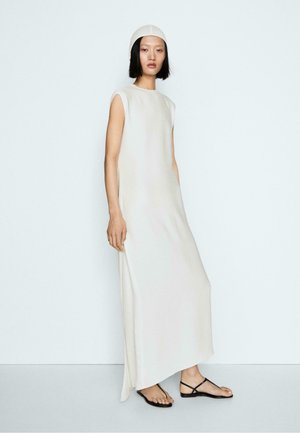 Donna in abito lungo bianco senza maniche e cappellino abbinato, indossa sandali neri bassi con cinturini, in piedi su sfondo bianco.