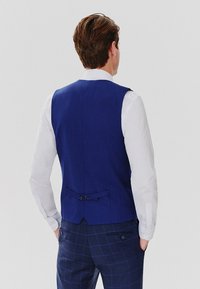 Granatowa kamizelka o teksturowym wykończeniu, z detalem w postaci ściągacza z tyłu i dwiema guzikami. Połączona z białą koszulą z długim rękawem.