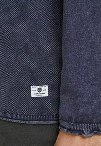 Tmavě modrá texturovaná látka s tkaným vzorem. Štítek u spodní části zní "Jack & Jones Premium, Quality Goods Co. ESTD 1975."