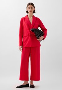 Woman wearing a red wide-leg pantsuit holding a black clutch bag, standing against a plain light gray background.
