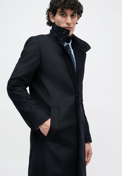 Cappotto lungo in misto lana blu navy con collo alto, chiusura frontale con bottoni e tasche laterali. Vestibilità adeguatamente sartoriale.