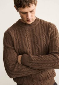 Un maglione marrone a coste lavorato a maglia presenta un design testurizzato con un collo rotondo a coste e maniche lunghe. Il materiale appare spesso e caldo.