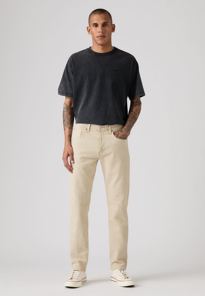 Uomo in piedi con le mani nelle tasche, indossa una maglietta nera sbiadita, jeans beige, sneakers bianche e mostra tatuaggi sulle braccia.