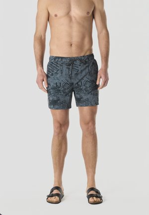 Hombre con pantalones cortos de baño de color gris oscuro con estampado y cordón ajustable, y sandalias negras, posando frente a un fondo liso y claro.