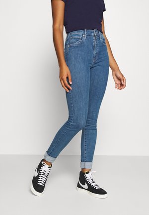 Person trägt hochgekrempelte blaue Skinny-Jeans, schwarz-weiße High-Top-Sneaker und ein dunkles kurzärmeliges Oberteil, steht vor einem einfarbigen Hintergrund.