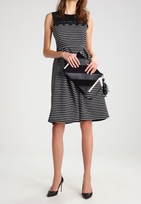 Kvinna i en svart och vit horisontellt randig ärmlös klänning som håller en svart handväska, bärande svarta högklackade skor, stående framför en vit bakgrund.