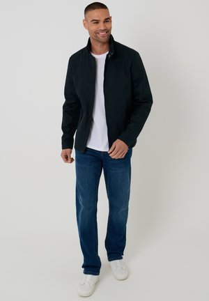 Man glimlachend, draagt een zwart jack met rits over een wit T-shirt, blauwe spijkerbroek en witte sneakers, staand tegen een effen lichte achtergrond.