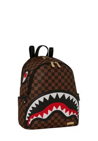 Zaino a scacchi marrone con un design a faccia di squalo, caratterizzato da dettagli rossi e bianchi, cerniere dorate e spallacci neri.