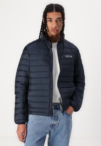 Jachetă puffer bleu marin cu un design matlasat orizontal, fermoar frontal și logo-ul Red Bull Racing pe pieptul stâng, purtată peste o cămașă deschisă.