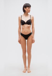 Γυναίκα που στέκεται ξυπόλυτη, φορώντας μαύρο σουτιέν και τα αντίστοιχα εσώρουχα, κοιτάζοντας προς τα δεξιά της, σε απλό φωτεινό φόντο.