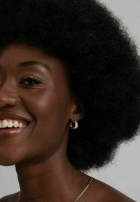 Nahaufnahme einer lächelnden Person mit Afrofrisur. Goldene Creolen, glatte Hautfarbe und eine entblößte Schulter mit einer subtilen Halskette.