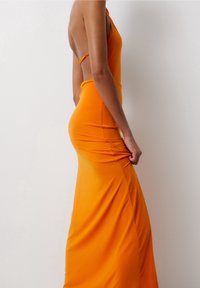 Robe orange moulante sans manches avec un dos haut et une coupe près du corps. Texture douce et lisse avec des détails froncés sur le côté.