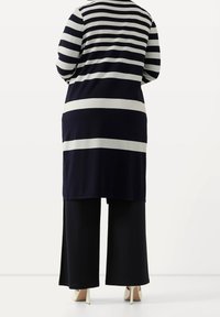 Dlouhý cardigan s námořnickými modrými a bílými pruhy, kombinovaný s širokými černými kalhotami a béžovými podpatky, pohled zezadu. Hladká textura a volný střih.
